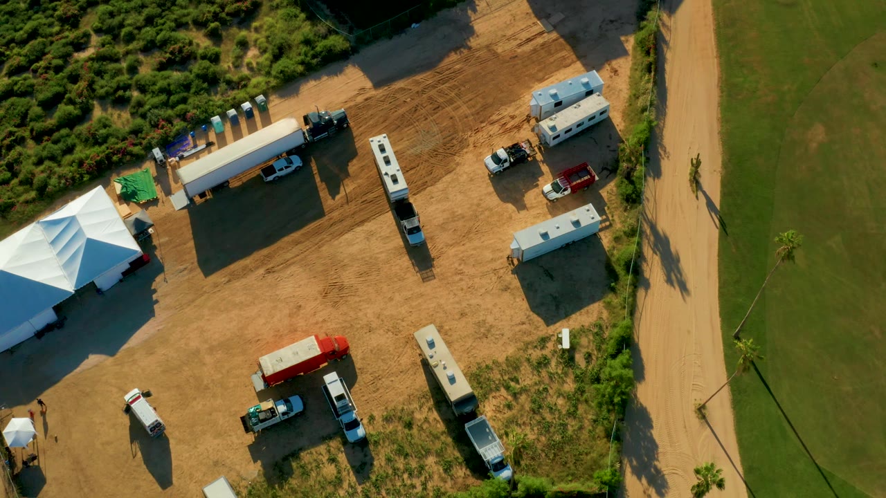 Top-down San Jose Del Cabo frame with the white tent cluster at left, RVs and trailers across the dirt lot, a red truck near the lower center, and the road and grass to the right, with no clearly visible people.