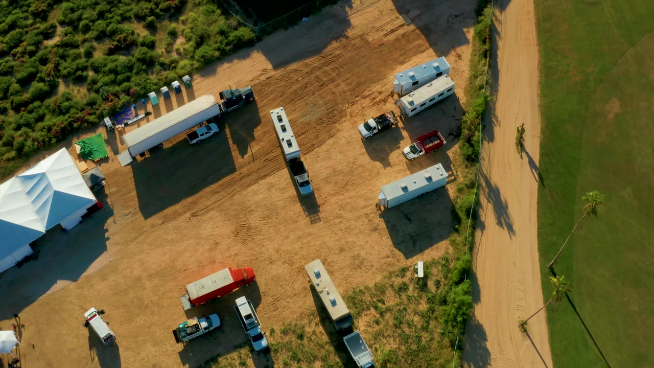 Aerial frame in San Jose Del Cabo with the white tents partly along the left edge, multiple trailers and vehicles across the dirt area, and the road running vertically on the right, with no clearly visible people.