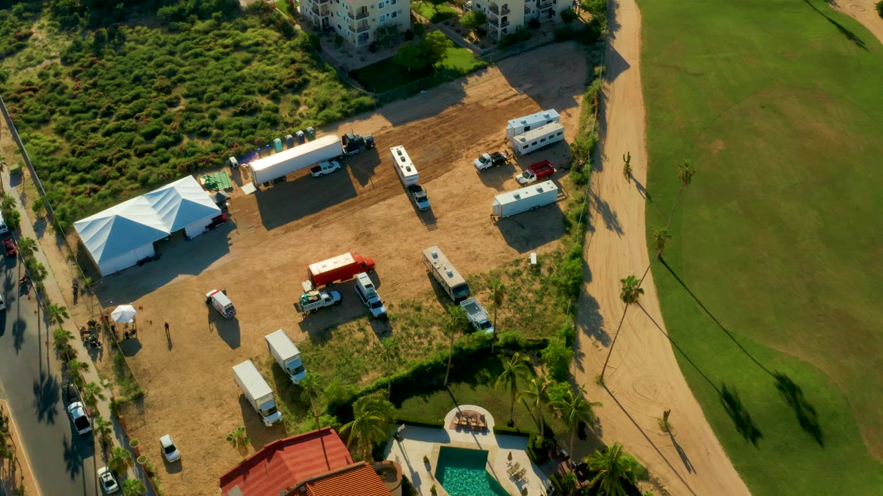 Overhead frame in San Jose del Cabo showing a dirt temporary site with a white tent, parked vehicles, a red object, a pool edge, green field, and no clearly visible people.