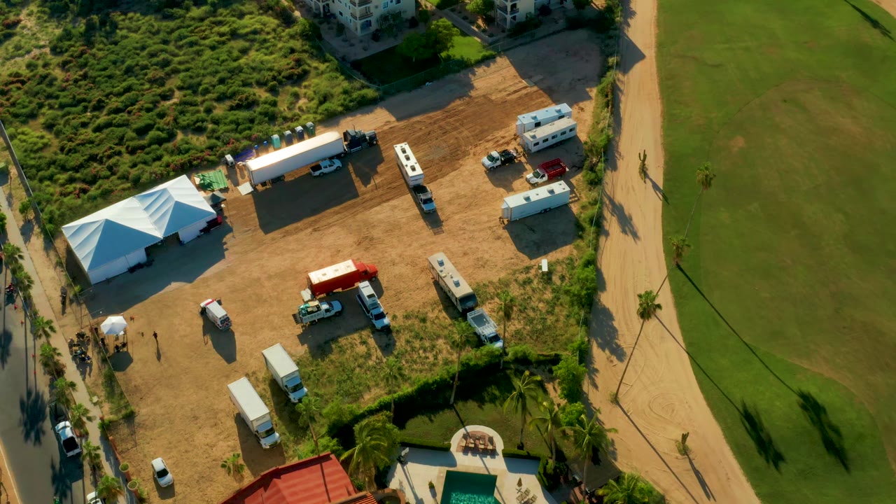 Overhead San Jose del Cabo frame showing the white truck near the center of the dirt site, with tents, trailers, parked vehicles, green turf, scrub vegetation, and no clearly visible people.