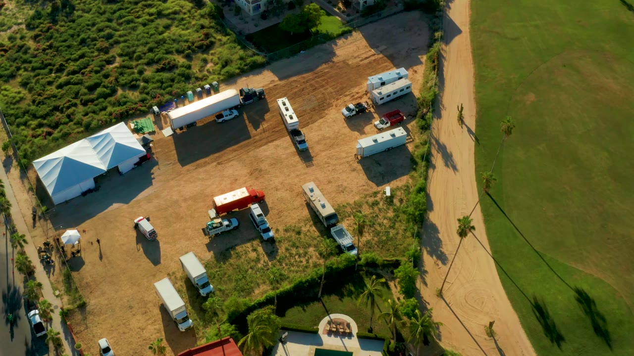 Drone view of a dirt lot with a tent, trucks, trailers, and green field edge.