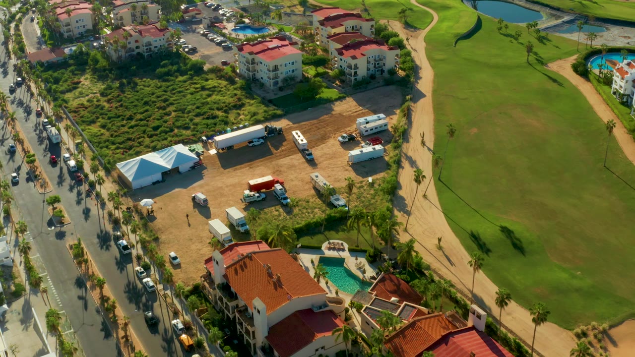 Aerial frame in San Jose Del Cabo with the road and moving cars along the left edge, red-roofed buildings near the top and bottom, a white tent in the dirt lot, and open green turf to the right, with no visible people.