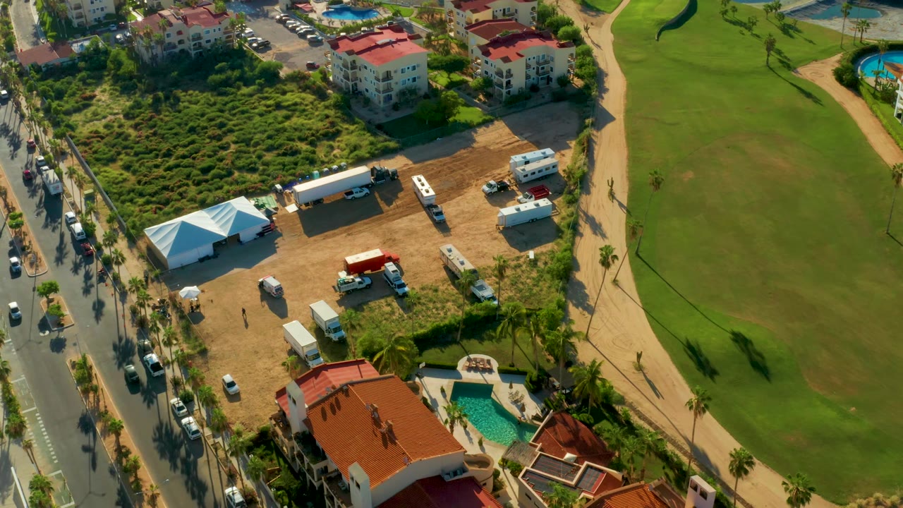 Aerial composition of a dirt lot, white tent, road, cars, and broad green field.