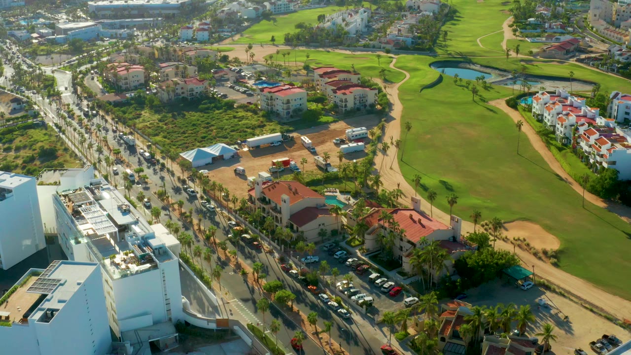 Aerial frame in San Jose Del Cabo with traffic on the left-side road, a broad green fairway, small pond, sand areas, red-roofed residences, and no visible people.