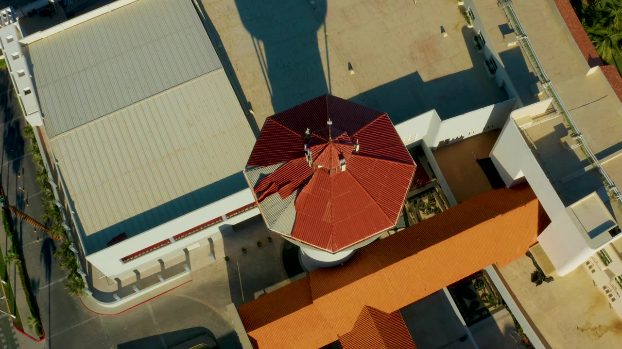 Top-down San Jose del Cabo frame showing the red octagonal roof near the right side, pale commercial roofs, a narrow street edge, warm orange roofing, and no visible people.