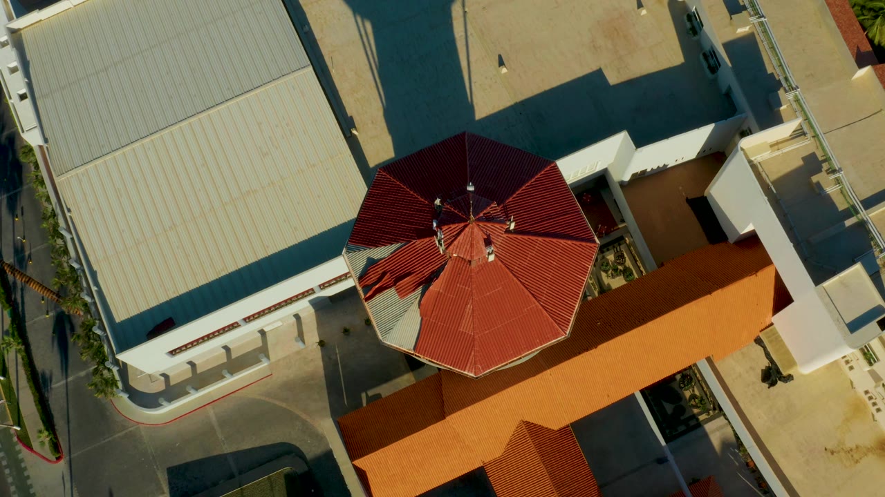 San Jose del Cabo aerial frame with the red octagonal roof lower in the composition, a large ribbed pale roof to the left, an orange roof section below, streets, and no visible people.