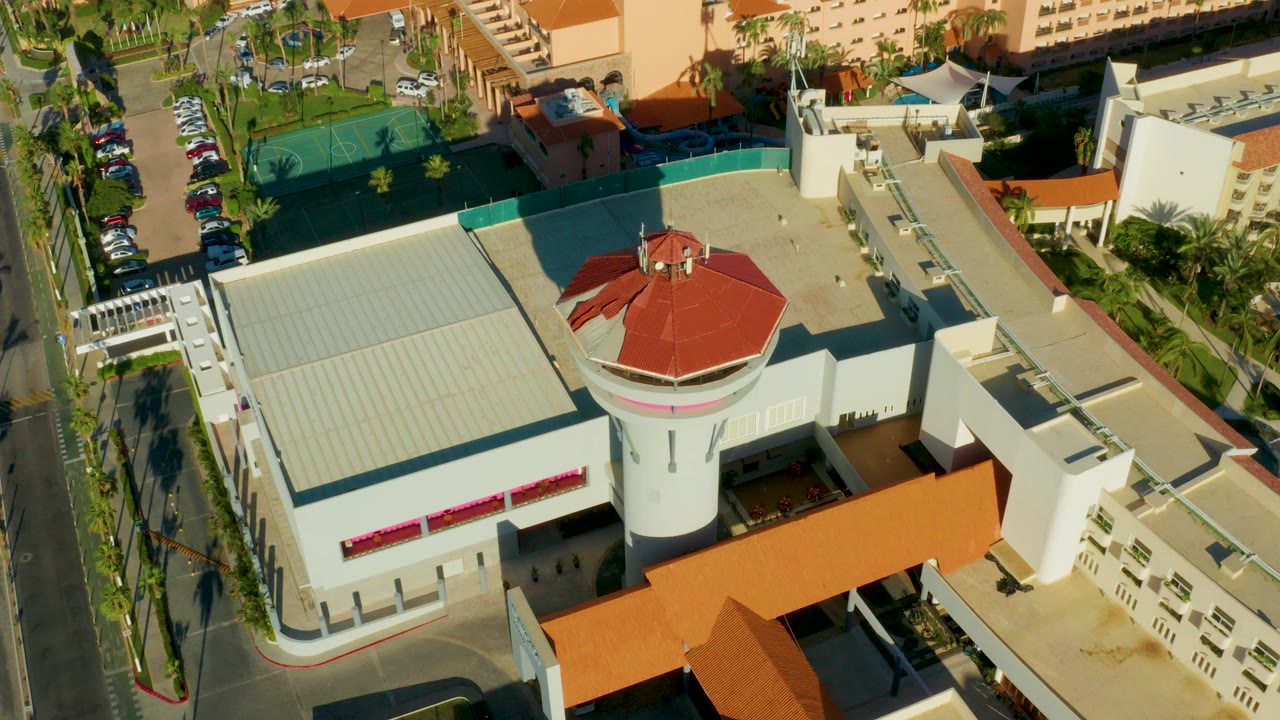 Overhead aerial frame in San Jose Del Cabo showing a white building complex with a red-tiled round tower, orange roof sections, green courts, parked cars, palm-lined streets, and no visible people.