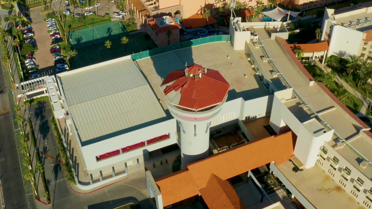 High-angle San Jose Del Cabo frame with the round red roof tower, white building walls, flat roof areas, nearby cars, green courts, palm trees, and no visible people.