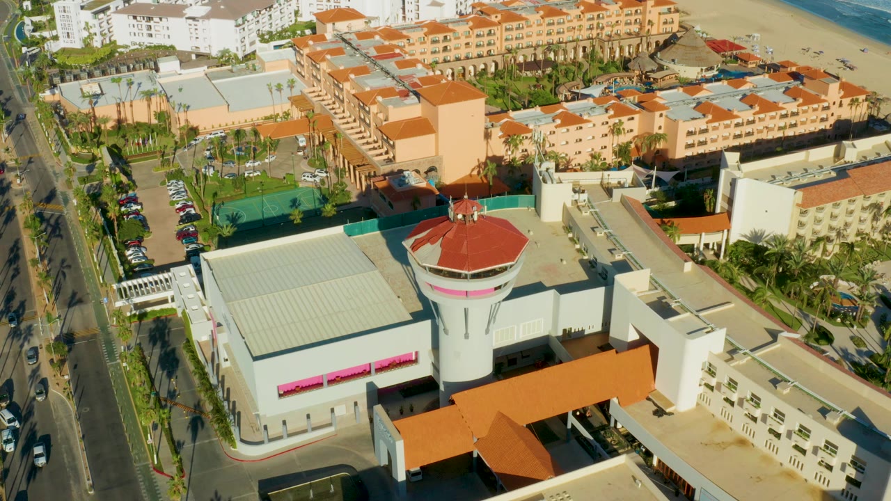 Opening aerial frame over San Jose del Cabo with a white round red-roofed tower, red-roofed buildings, a palm-lined road, parked cars, sandy beach, ocean, and no visible people.