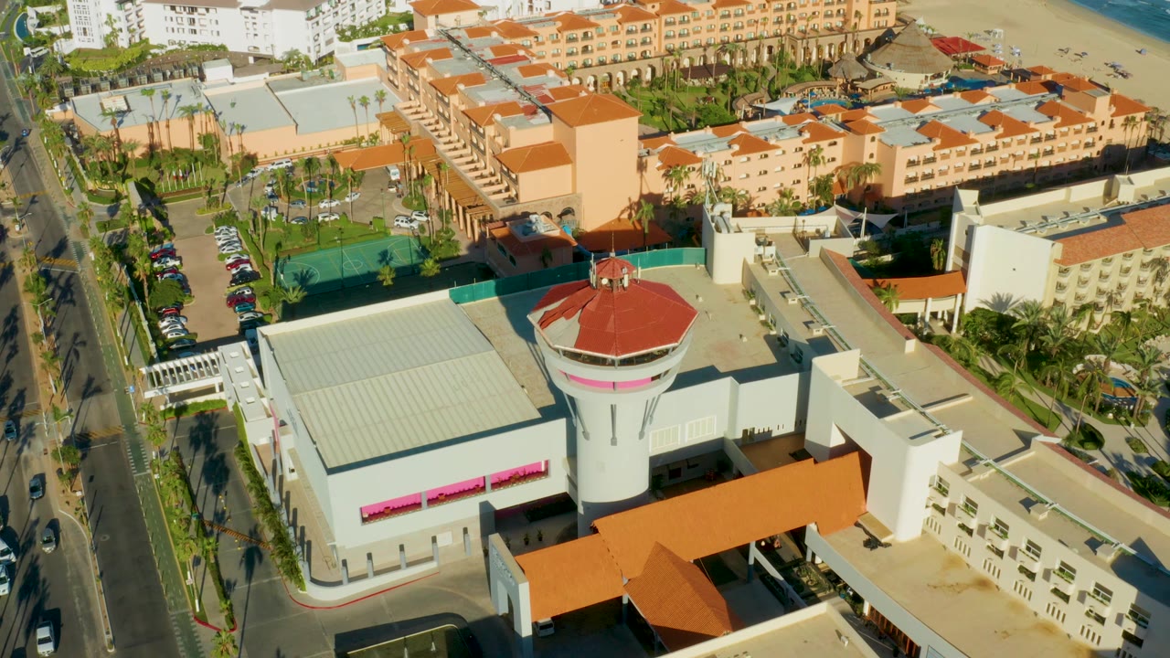 Aerial San Jose del Cabo frame with the round red-roofed building slightly right of center, a straight road with cars along the left edge, clustered resort-style buildings, beach, ocean, and no visible people.