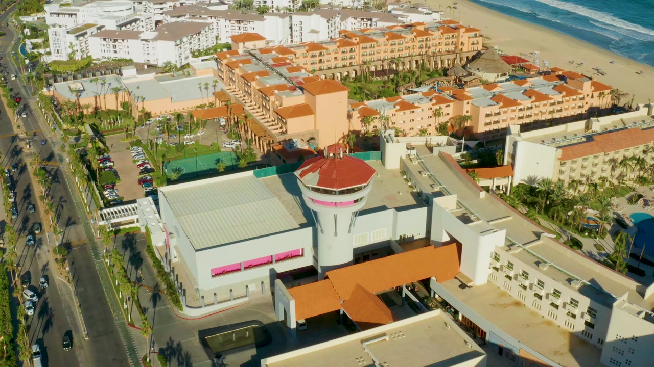 Aerial opening frame over San Jose del Cabo with white and tan hotel buildings, orange roofs, road traffic at left, sandy beach and blue ocean at top, and no visible people.