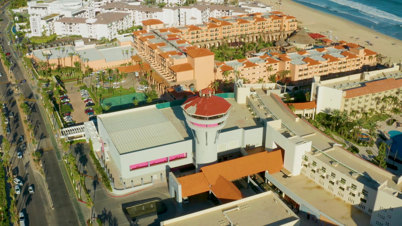 Drone view of hotel roofs, palm trees, road vehicles, beach, and ocean.
