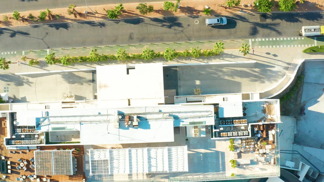 Top-down view in San Jose del Cabo showing a white flat-roofed building beside a divided street with parked and moving cars, trees, and no visible people.