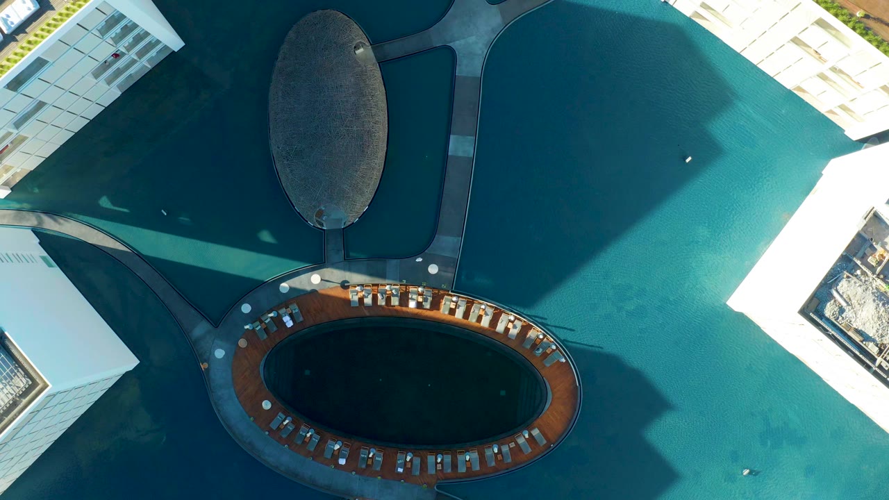 Overhead frame from San Jose Del Cabo showing turquoise water around white modern buildings, curved concrete paths, a dark oval plaza feature, and no visible people.