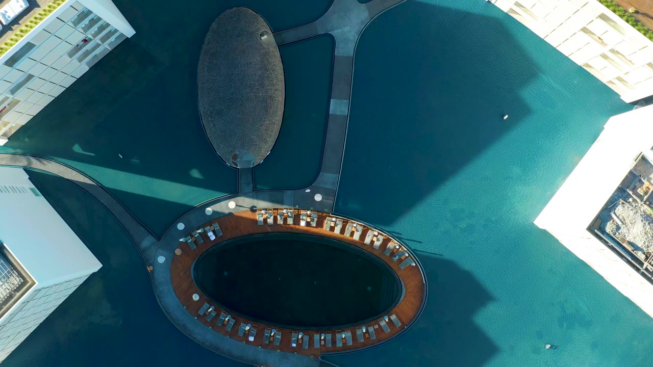 Aerial frame from San Jose Del Cabo with the dark central oval feature near the lower edge, white building facades, turquoise water, and no visible people.