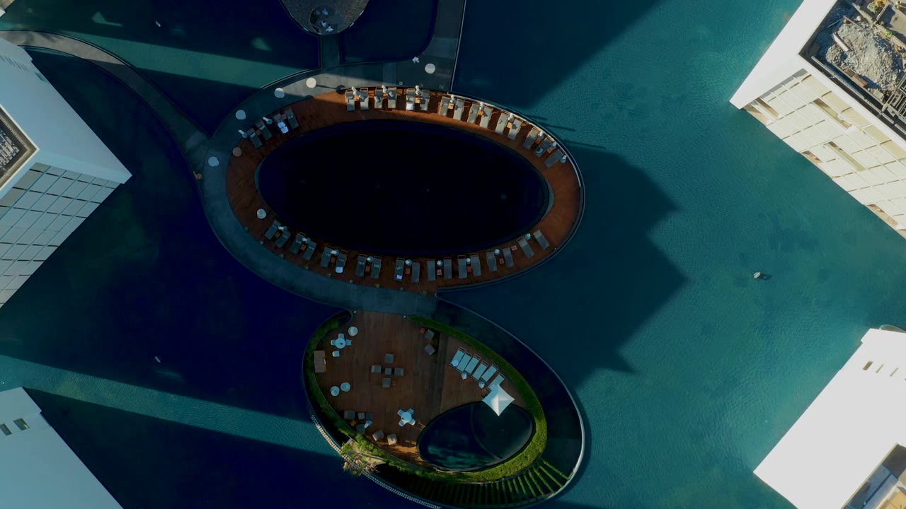 Aerial frame over San Jose Del Cabo with the larger dark circular pool near the top, a smaller rounded deck below, bright building edges, teal water, and no visible people.