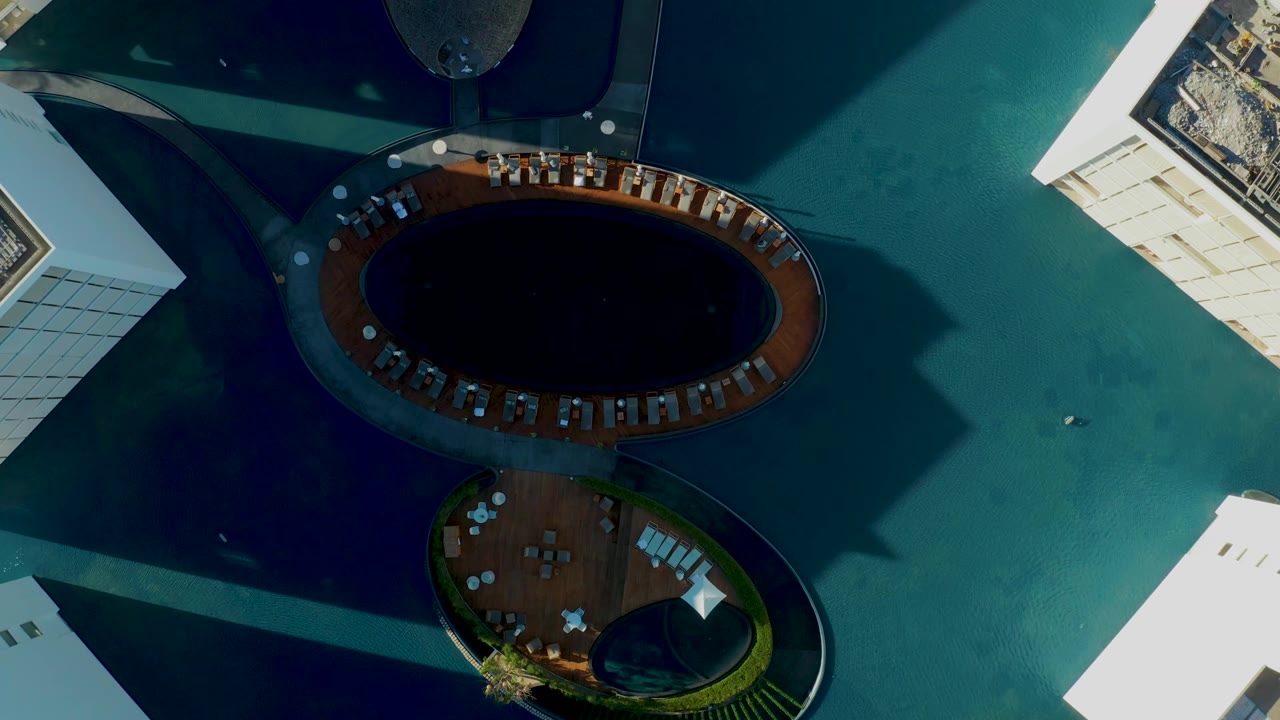 Top-down San Jose Del Cabo frame with the dark pool shifted higher in view, the curved deck below it, pale building blocks at the edges, blue-green water, and no visible people.