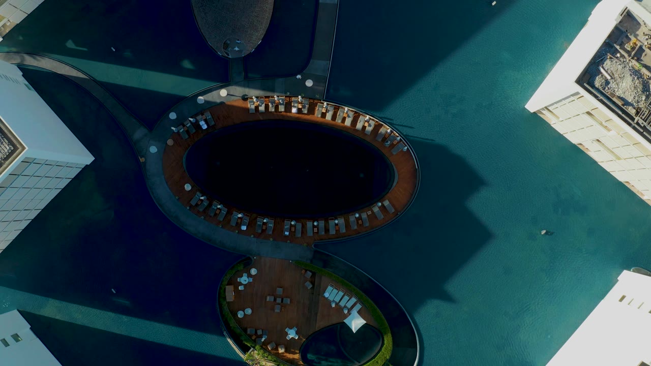 Mid-clip overhead view in San Jose Del Cabo showing an oval dark pool ringed by lounge chairs, a rounded lower deck, angular building shadows, blue-green water, and no visible people.