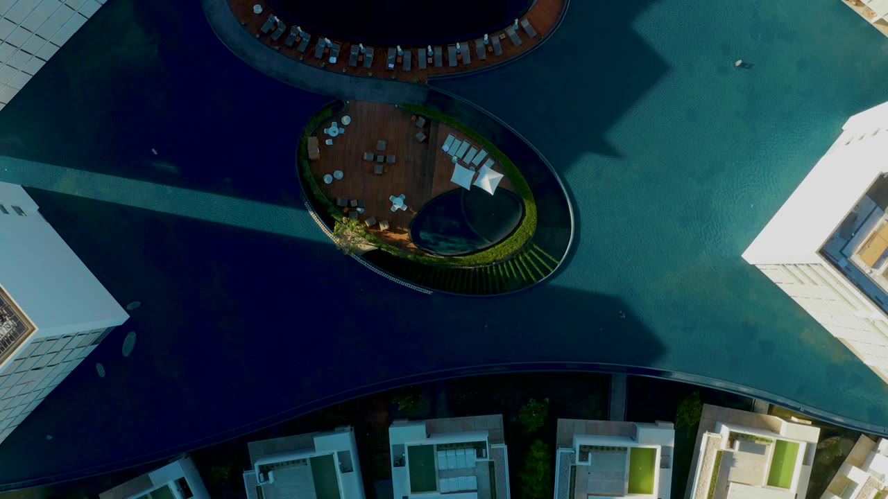 Aerial overhead frame in San Jose Del Cabo showing the circular deck lower in frame, the upper pool rim near the top, white building edges, blue-green water, and no visible people.