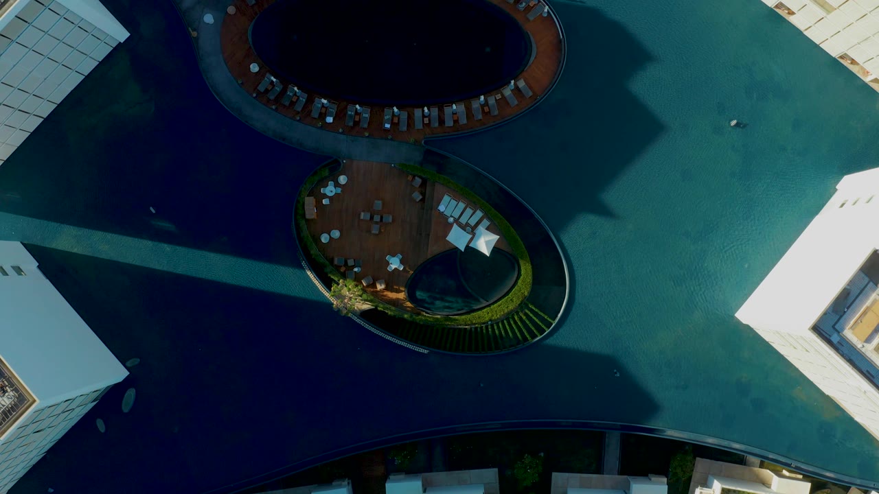 Overhead frame from San Jose Del Cabo showing the upper pool as a dark oval, the lower circular deck with tables, surrounding blue-green water, white architecture, and no visible people.