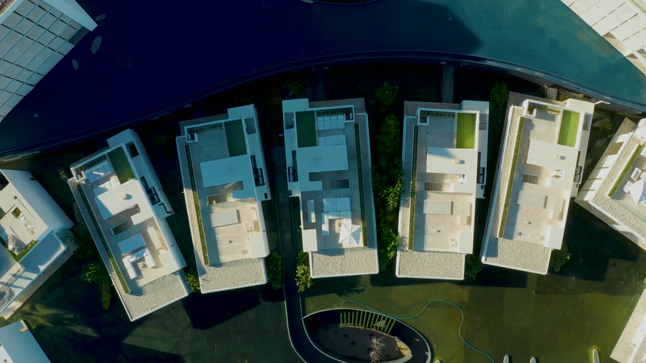 Top-down aerial frame in San Jose Del Cabo showing white apartment buildings, a curved dark pool, green lawn, paved areas, and no visible people.