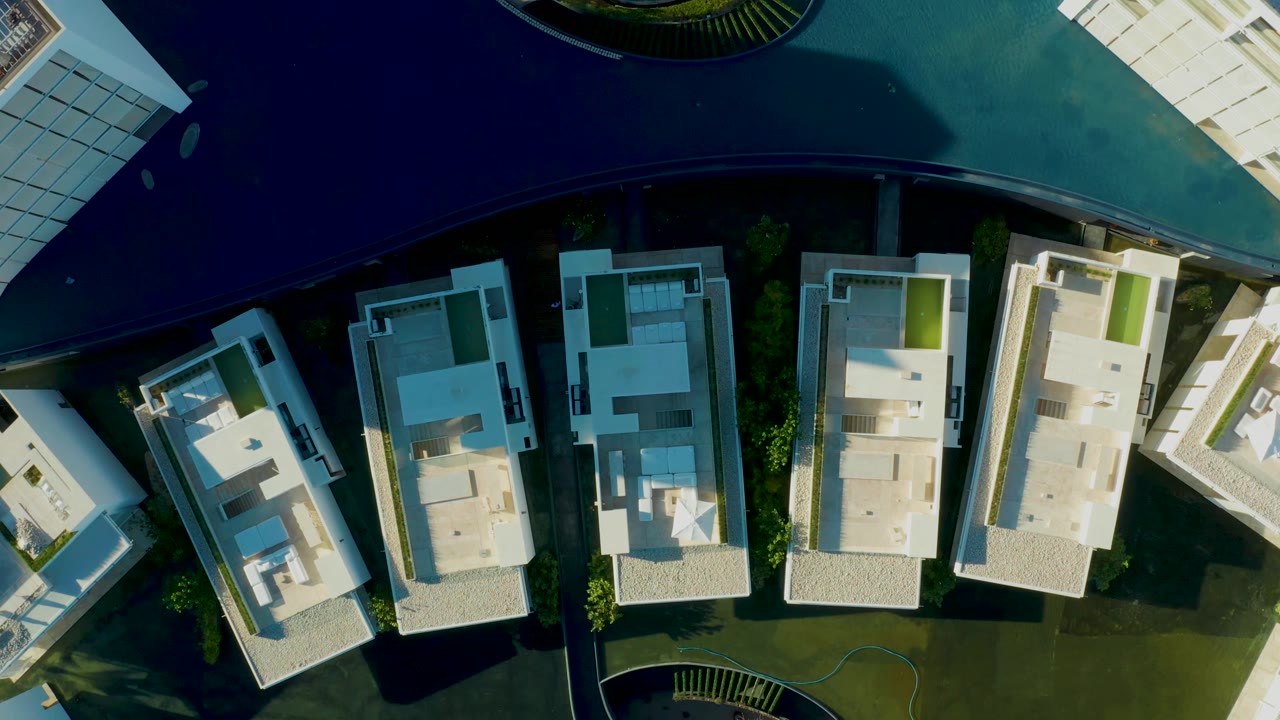 Top-down San Jose Del Cabo frame showing more of the apartment roofs, dark curved pool water, clipped lawn edges, paved paths, and no visible people.