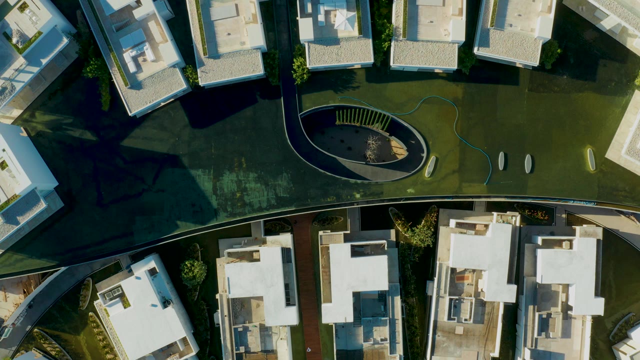 Opening overhead frame of a modern residential complex in San Jose Del Cabo, with white flat-roofed buildings, green lawn, an oval landscaped area, long shadows, and no visible people.