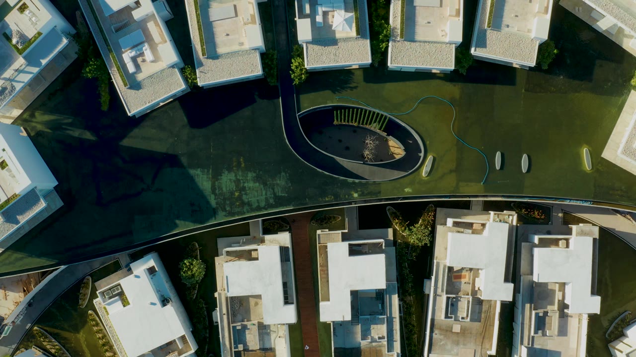 Second overhead frame in San Jose Del Cabo showing more of the curved circulation path between white residential buildings, green lawn, shaded pavement, and no visible people.