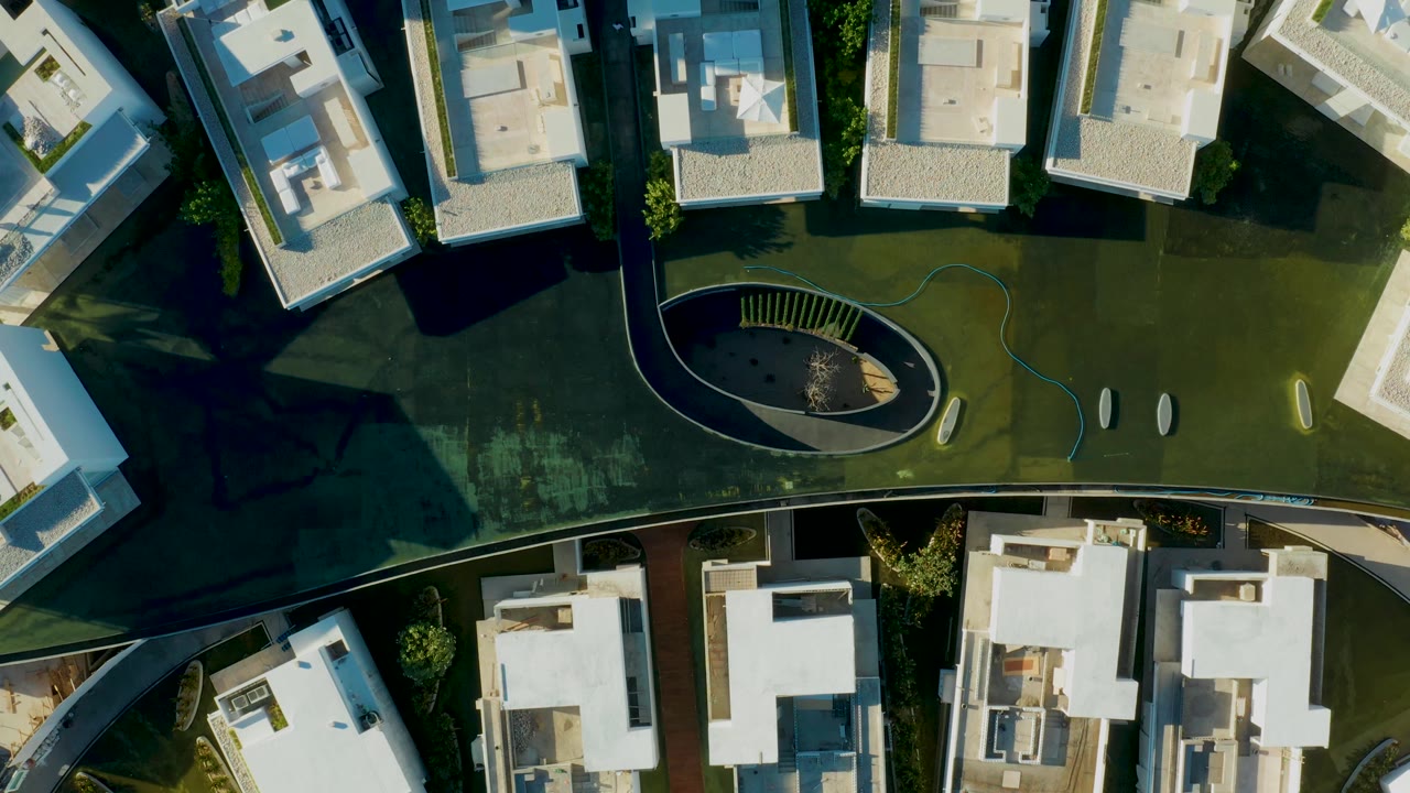 Third frame of the San Jose Del Cabo aerial view, with white buildings on both sides of a dark curved path, a green courtyard surface, an oval feature near center, and no visible people.