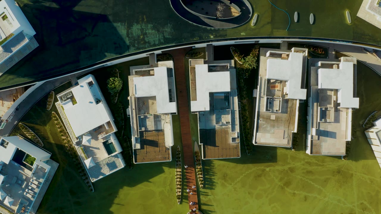 Overhead San Jose Del Cabo frame showing modern homes, rectangular rooftops, green grass, a dark paved section, and no visible people.