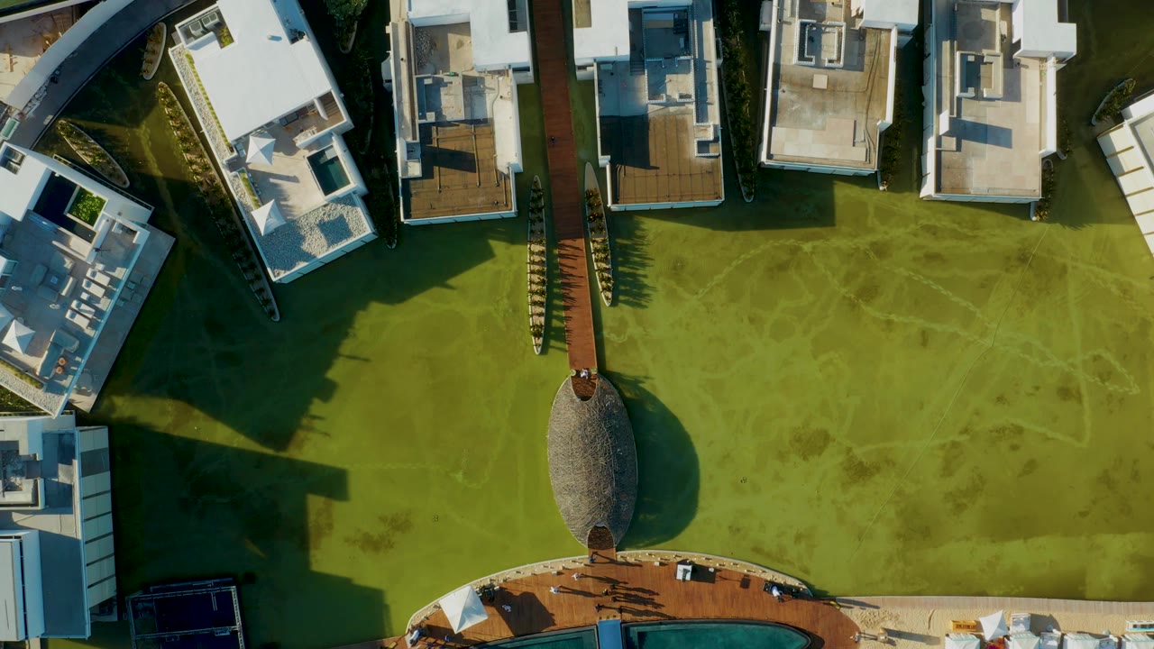 Aerial overhead frame in San Jose del Cabo where the pool edge is nearly out of view, with the central path, oval thatched feature, white roofs, green lawn, and no visible people.