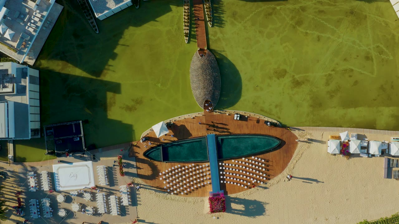 Top-down aerial frame in San Jose Del Cabo showing a dark pool in a wood deck, rows of loungers on sand, green water or turf above, resort buildings at the edges, and small people visible near the poolside.