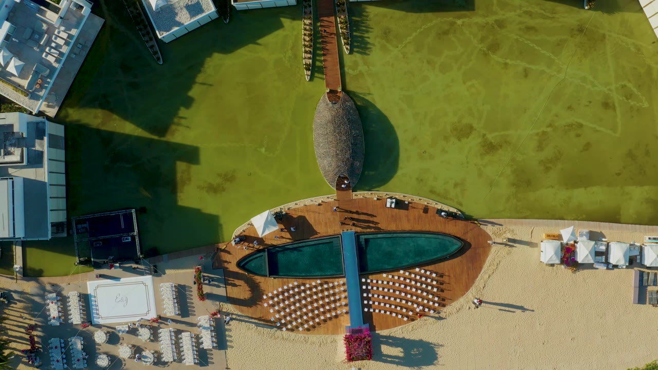 At 0:02, the overhead San Jose Del Cabo view shows the pool and deck shifted slightly within the grid cell, with lounge chairs on sand, buildings along the sides, and small people still visible near the resort area.