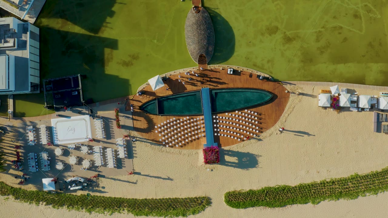 At 0:02, the overhead San Jose del Cabo view shows the pool deck between green lawn and sand, with white lounge furniture, umbrellas, cabanas, and tiny visible people.