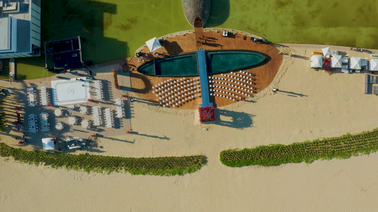 Overhead view in San Jose Del Cabo showing a narrow pool in a wooden deck, rows of lounge chairs on sand, white umbrellas, green lawn, and small visible people near the seating.