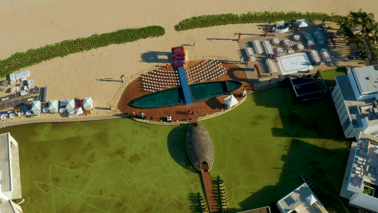 Overhead view of a San Jose del Cabo beach resort with sand at the top, an oval pool and lounge chairs in the center, green lawn below, and no visible people.