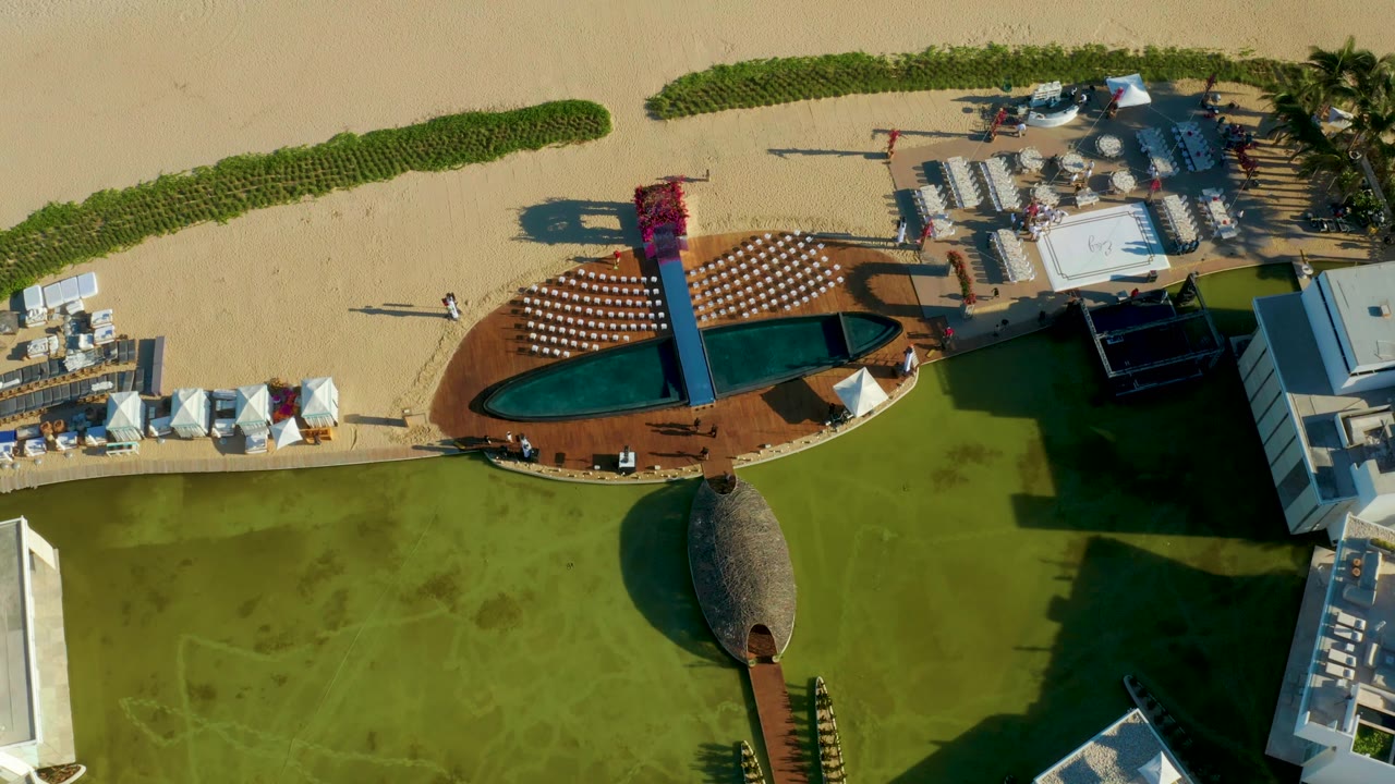 Top-down San Jose del Cabo resort view with the red slide structure crossing the pool area, rows of loungers on the deck, sandy beach above, and no visible people.