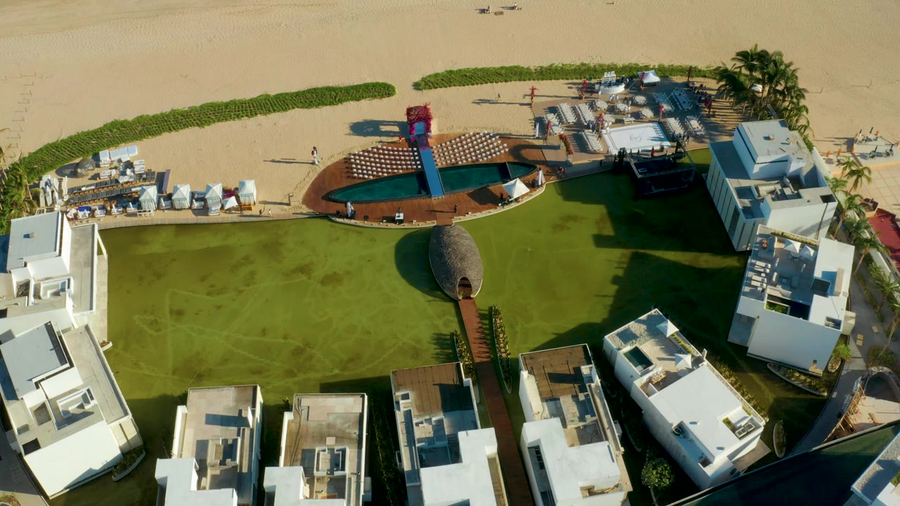 Overhead frame of a San Jose Del Cabo beach resort with a green lawn, small pool, brown deck, white buildings, sand, and no visible people.