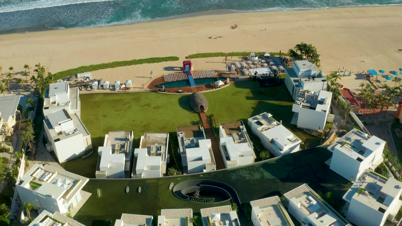 Aerial view in San Jose del Cabo showing white resort buildings around a green lawn and curved pool area, with sandy beach and blue-green surf behind them and no visible people.