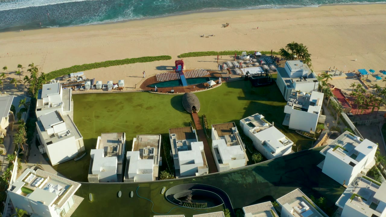 Drone frame over San Jose del Cabo resort grounds, with foreground white rooftops, a central green lawn, a small curved pool area, beach sand, and visible ocean break, with no visible people.