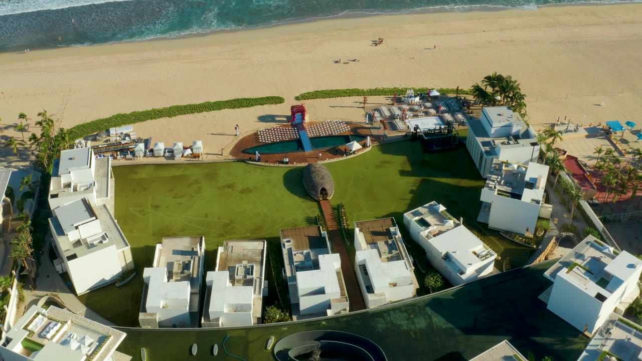 Aerial resort grounds with white structures, green lawn, pool deck, beach sand, and waves.