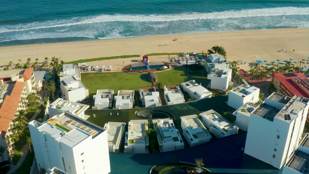 San Jose Del Cabo resort roofs sit below a sandy beach and active surf line, with blue umbrellas near the palms and no visible people.