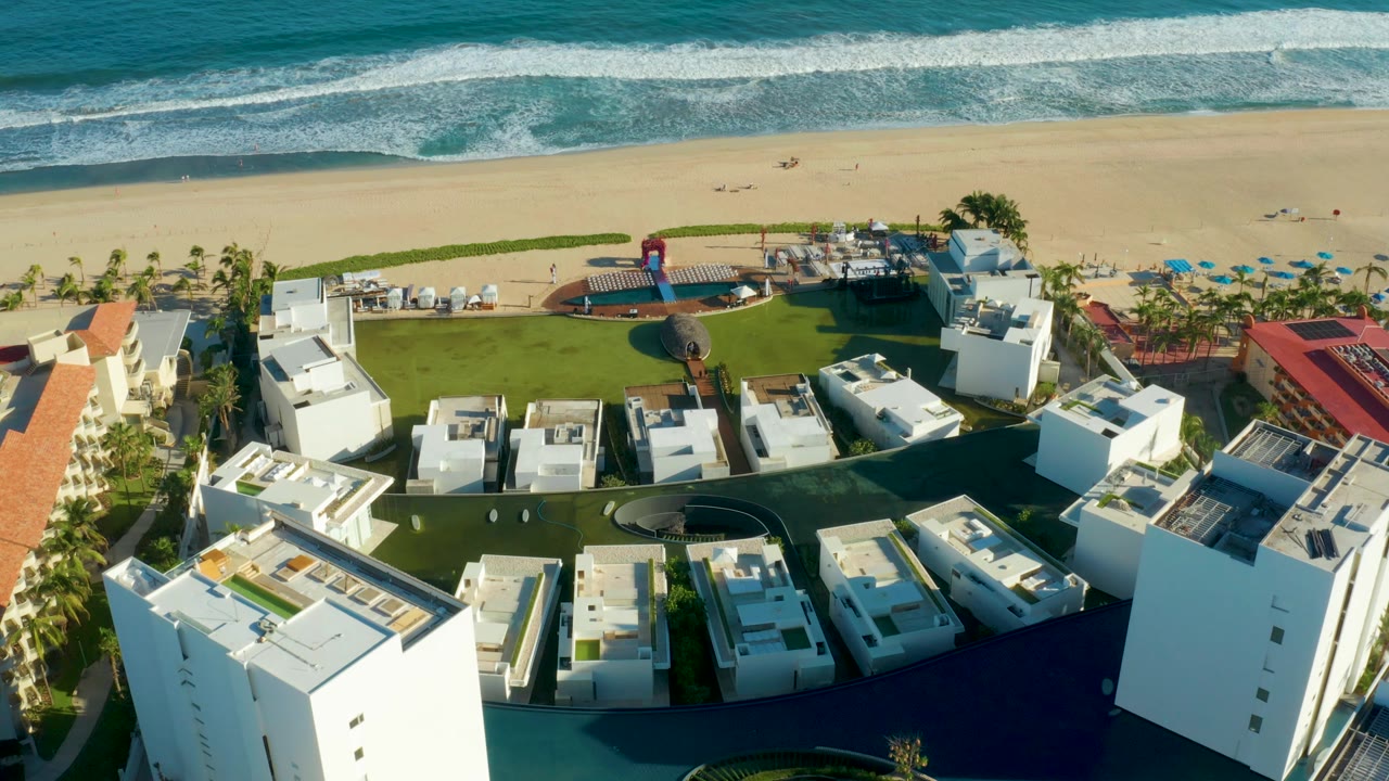 The aerial view of the San Jose Del Cabo resort shows white structures clustered around lawns, with the beach and foamy ocean waves across the background and no visible people.