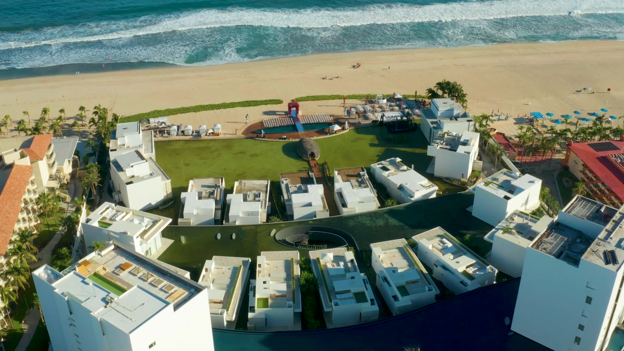 Aerial beach resort frame with white buildings, lawn, palms, and surf.