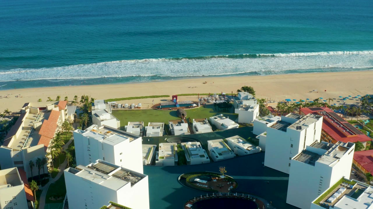 Aerial view of white resort buildings beside a beach and turquoise ocean.