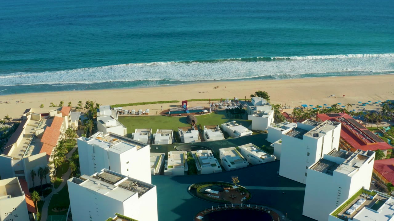 Aerial view in San Jose Del Cabo with resort roofs and white walls below, the swimming pool near the center, beach sand above it, and a wide band of ocean surf with no visible people.