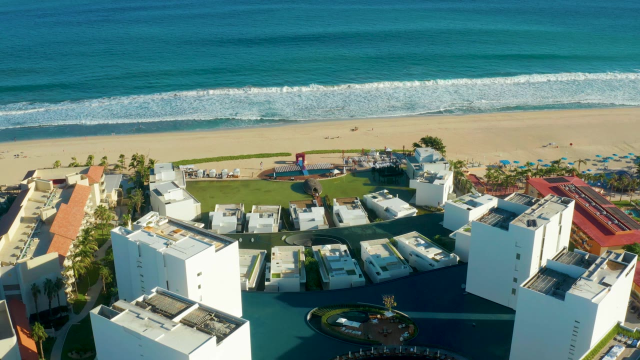 Mid-clip aerial over San Jose Del Cabo resort buildings, with the pool and dark courtyard in the lower area, palms along the beach edge, pale sand, and blue-green ocean waves with no visible people.