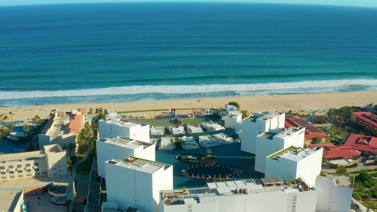 San Jose del Cabo frame showing white rooftops and a dark circular pool below, red-roofed buildings to the right, pale sand and turquoise ocean bands behind, with no visible people.