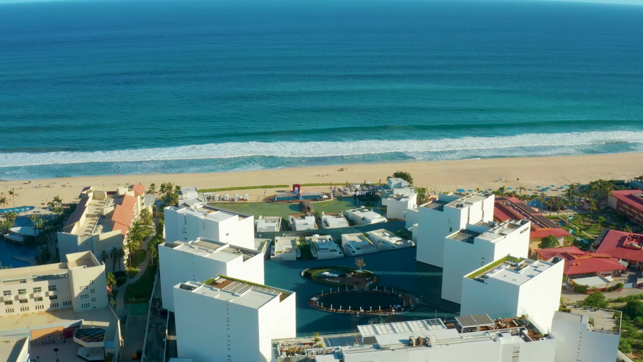 San Jose del Cabo aerial still with bright white building blocks, a central pool area, red roofs along the right edge, a narrow beach strip, and blue-green ocean water, with no visible people.