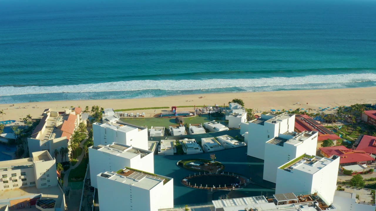 Drone-height view over white buildings toward a sandy beach and rolling surf.